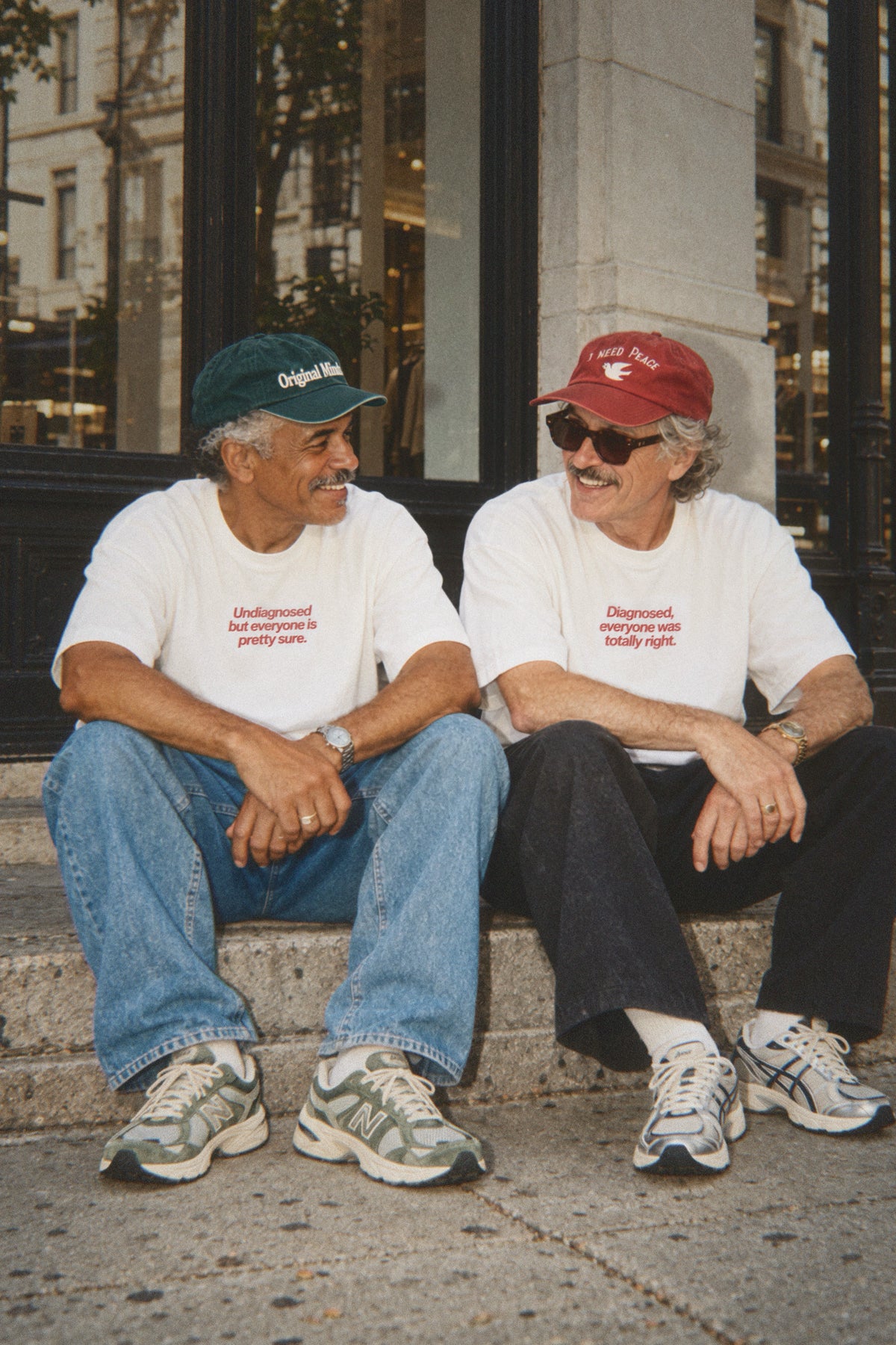 Two men in caps and Original Minds T-shirts sitting on a doorstep, smiling at each other